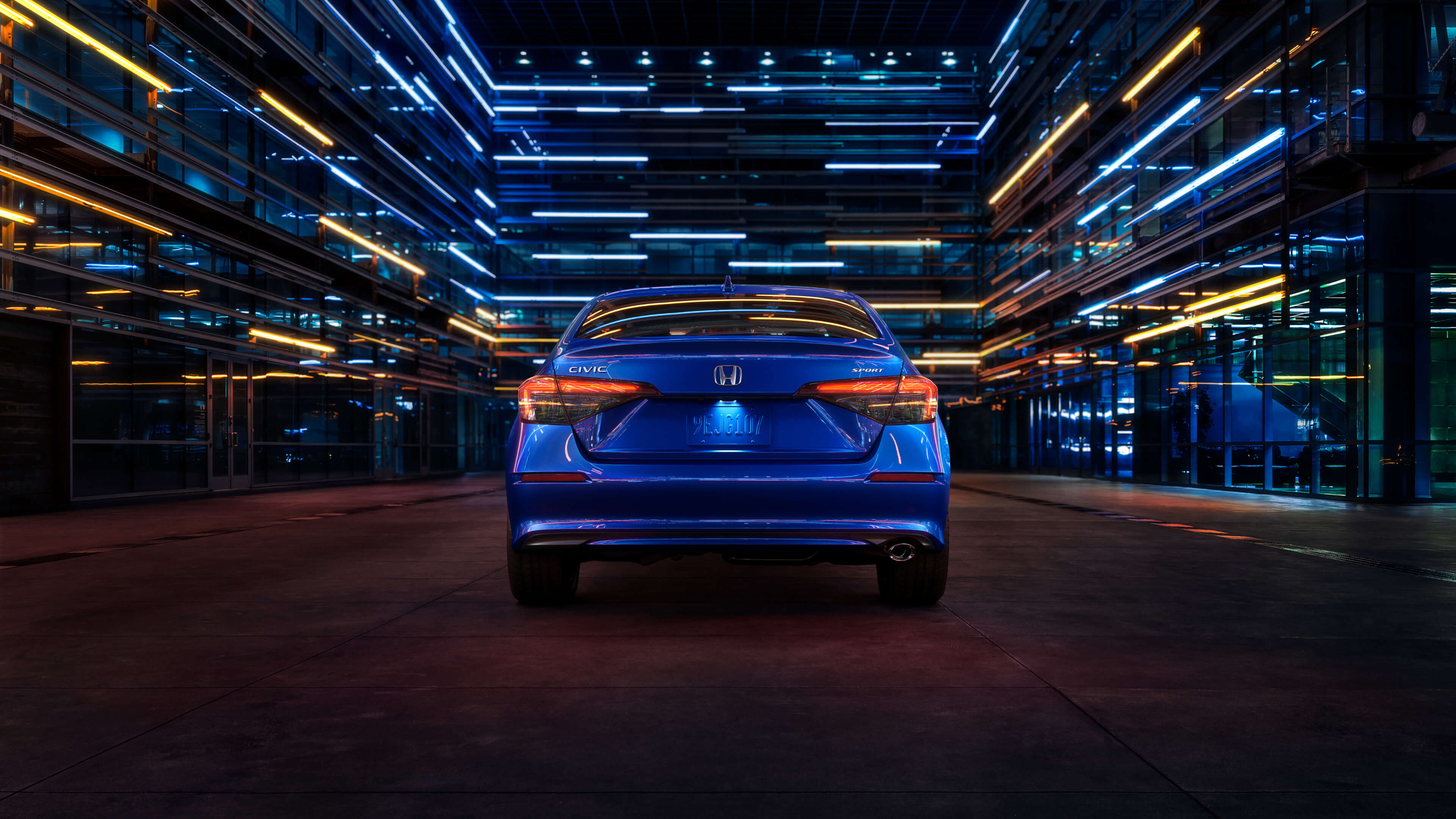 Vue arrière d’une Honda Civic 2022 bleue circulant de nuit dans une rue urbaine éclairée par des lumières bleues et blanches provenant des bâtiments environnants.